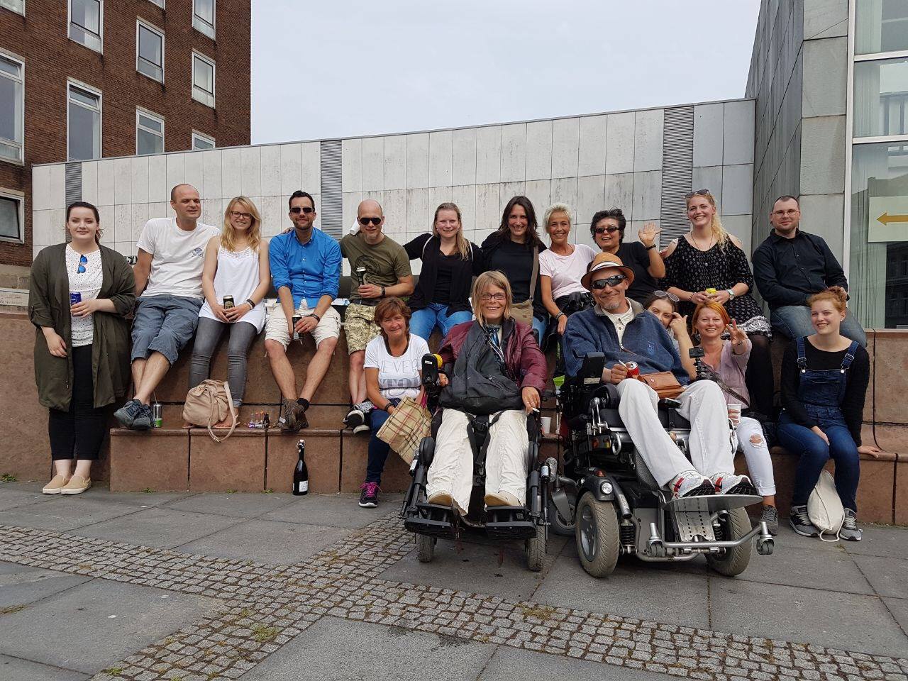 Eine Gruppe von Menschen, darunter zwei im Rollstuhl, posiert im Freien auf einer Treppe vor einem Gebäude.