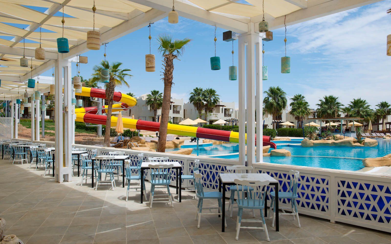 Resort dining area overlooking the pool, surrounded by palm trees and sun loungers.