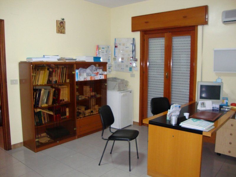 Interno dello studio medico con scrivania, librerie, minifrigo e doppie porte chiuse in legno.