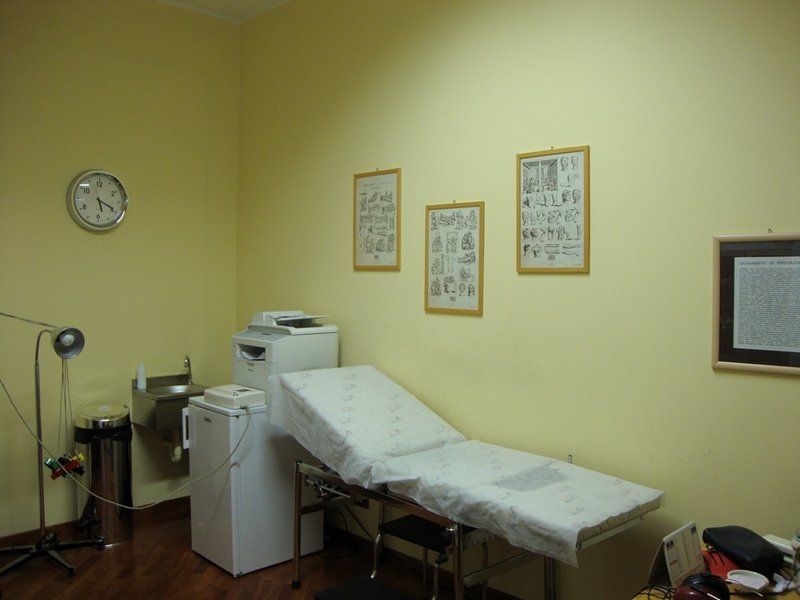 Sala visite mediche con letto, attrezzatura, diagrammi incorniciati e orologio. Pareti gialle.