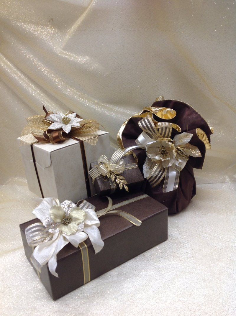 Tre scatole regalo nei toni del marrone e del bianco, decorate con eleganti fiori in tessuto e nastri su una superficie scintillante.