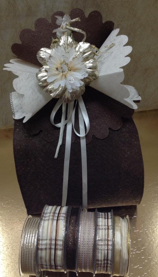 Una scatola regalo marrone sormontata da un fiocco bianco smerlato e da decorazioni floreali, posizionata davanti a diversi rotoli di nastro.