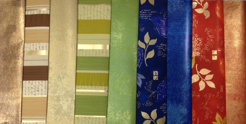 Nove rotoli di carta da parati in vari motivi e colori, tra cui marrone, verde, blu, rosso e tonalità metalliche con effetto materico.