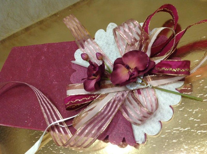 Composizione decorativa di fiori e nastri color bordeaux su una base bianca smerlata, su sfondo dorato.
