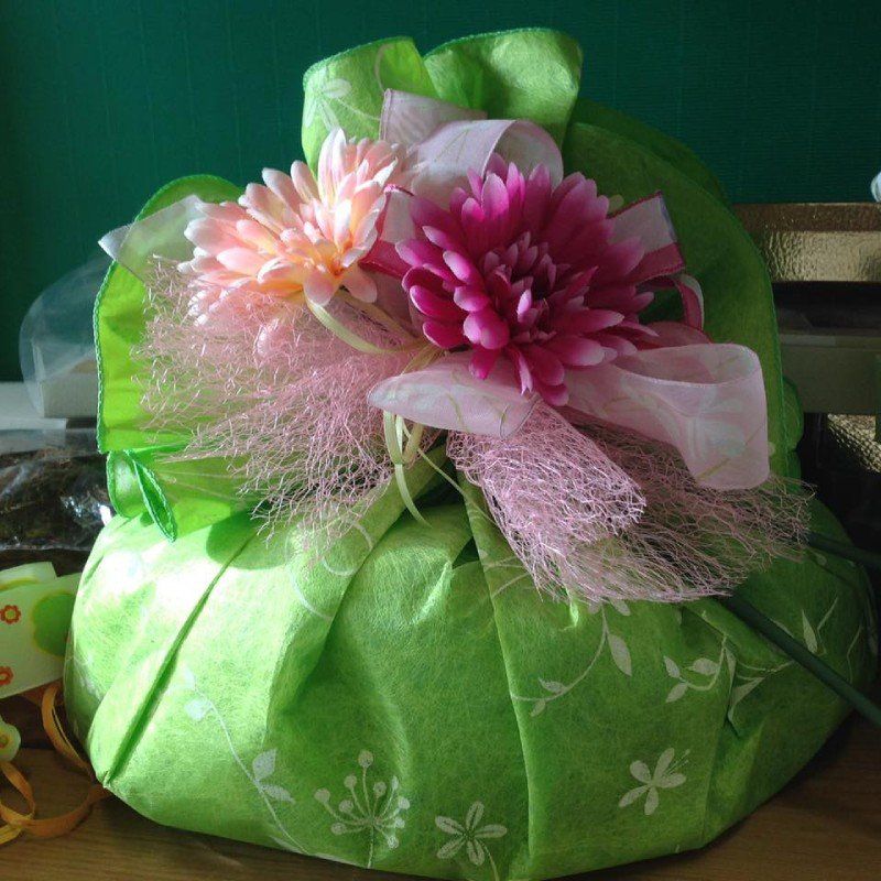 Un regalo rotondo avvolto in un tessuto verde chiaro con un motivo floreale bianco, decorato con fiori rosa e nastri nella parte superiore.