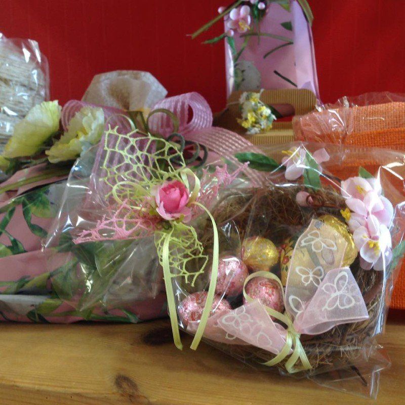 Cesti regalo con uova di cioccolato, fiori decorativi e nastri, esposti su una superficie di legno.