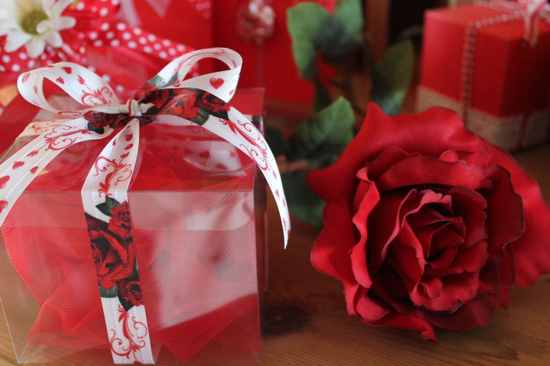 Una confezione regalo avvolta da un nastro bianco decorato con cuori e rose rosse, posizionata accanto a una grande rosa rosso intenso.