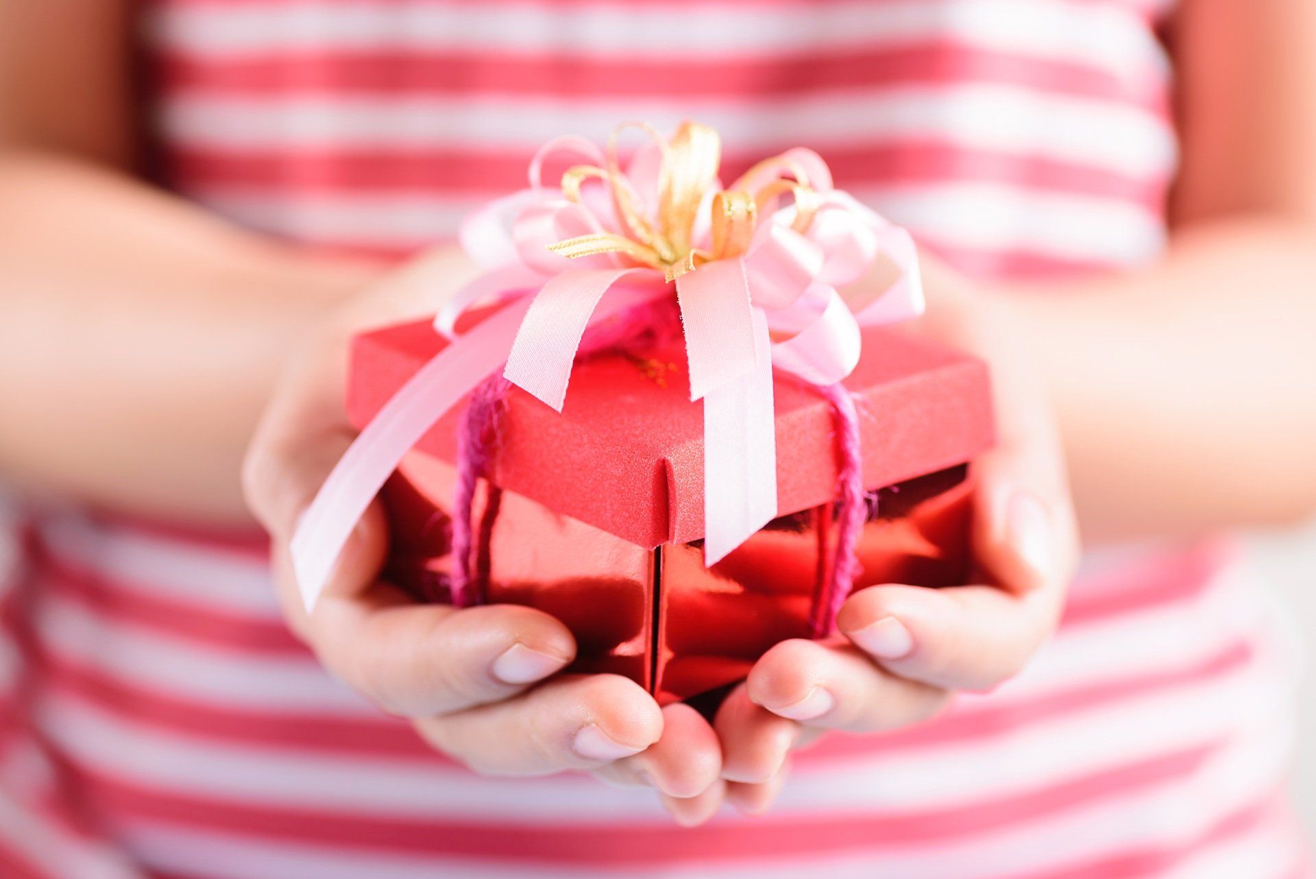 Due mani reggono una piccola scatola regalo rossa, legata con un nastro decorativo rosa.
