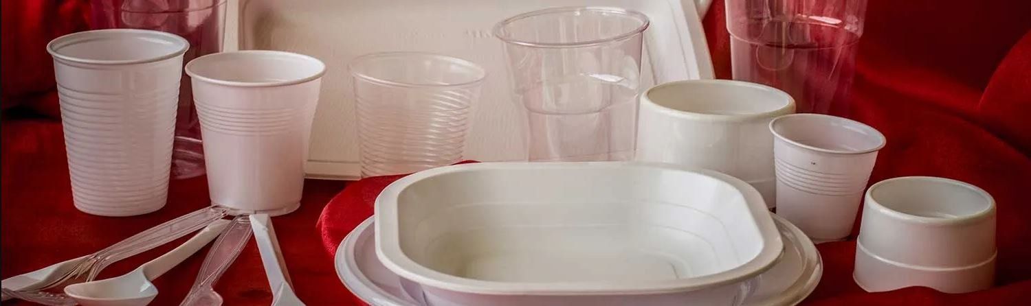 Vari tipi di bicchieri, ciotole e posate di plastica bianca usa e getta, disposti su una superficie rossa.