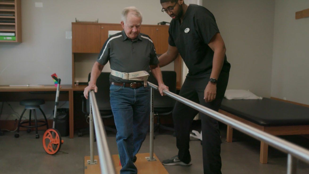 A man is helping an older man walk on parallel bars.