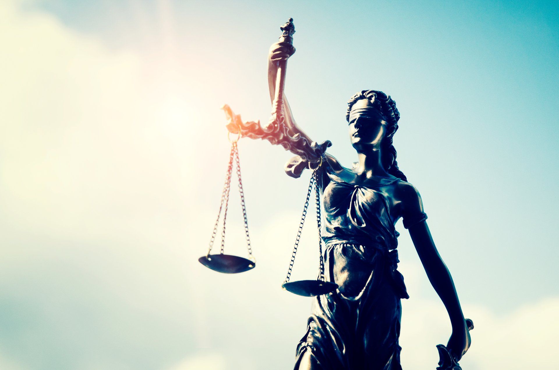 Statue of Lady Justice holding scales, against a bright blue sky with sunburst.
