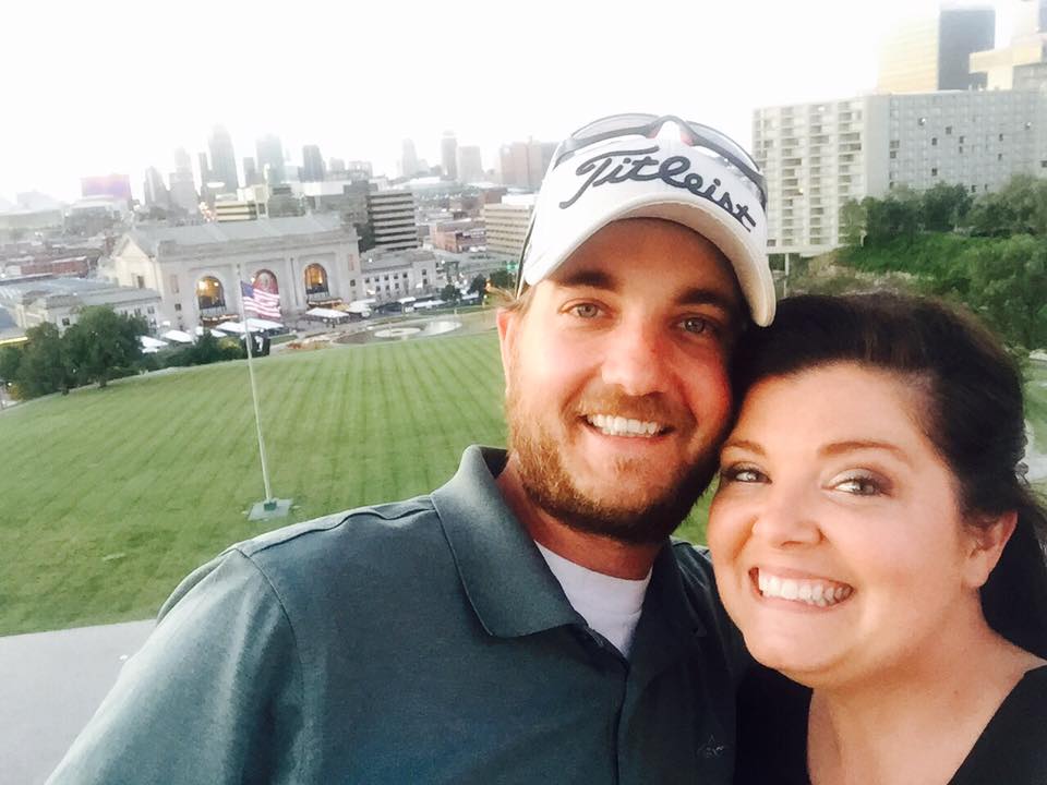 Smiling couple posing for a selfie at a stadium with a city skyline behind them
