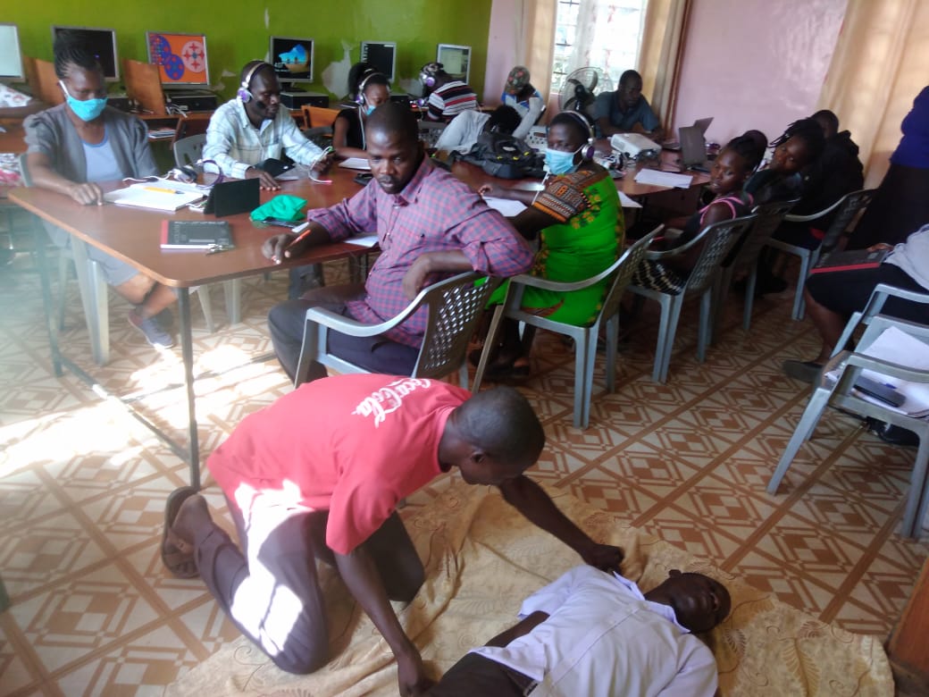 A group of people are sitting at tables and chairs in a room while a man is kneeling down to help another man