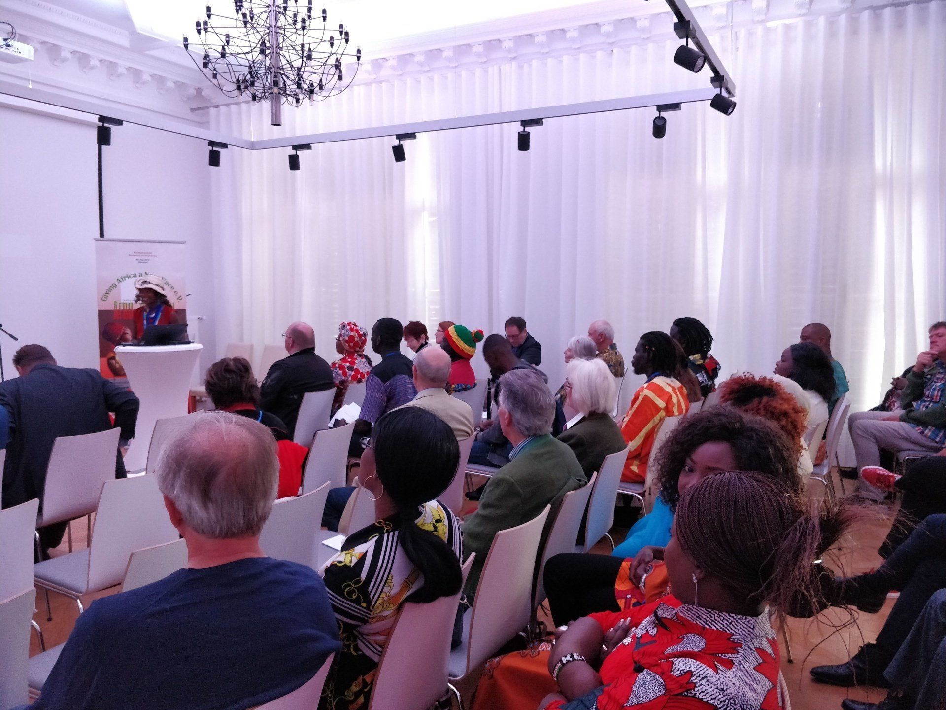 A group of people are sitting in chairs in a room with white curtains.