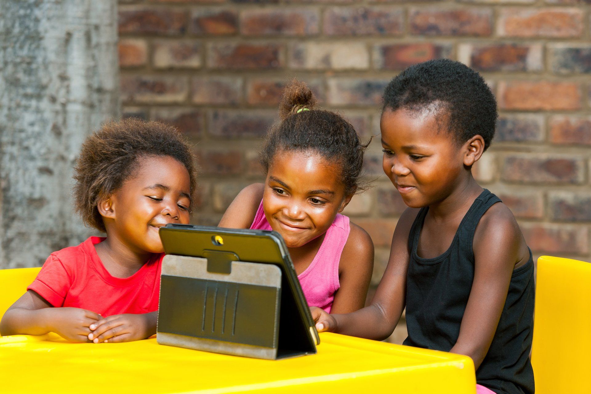 Drei Kinder sitzen an einem Tisch und schauen auf ein Tablet.