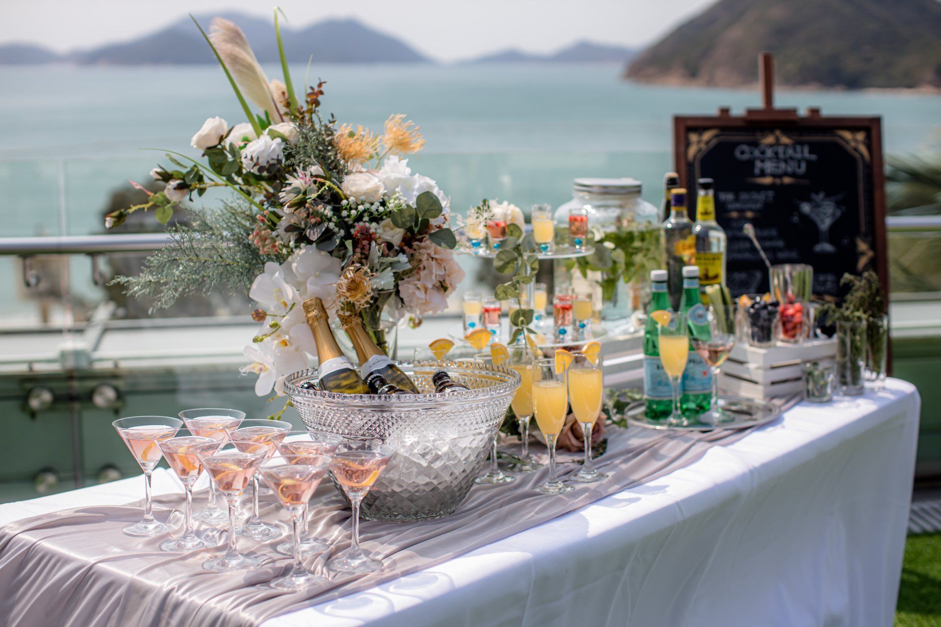 Cocktail bar with rose champagne, mimosa drinks, floral arrangement, and sea view background.