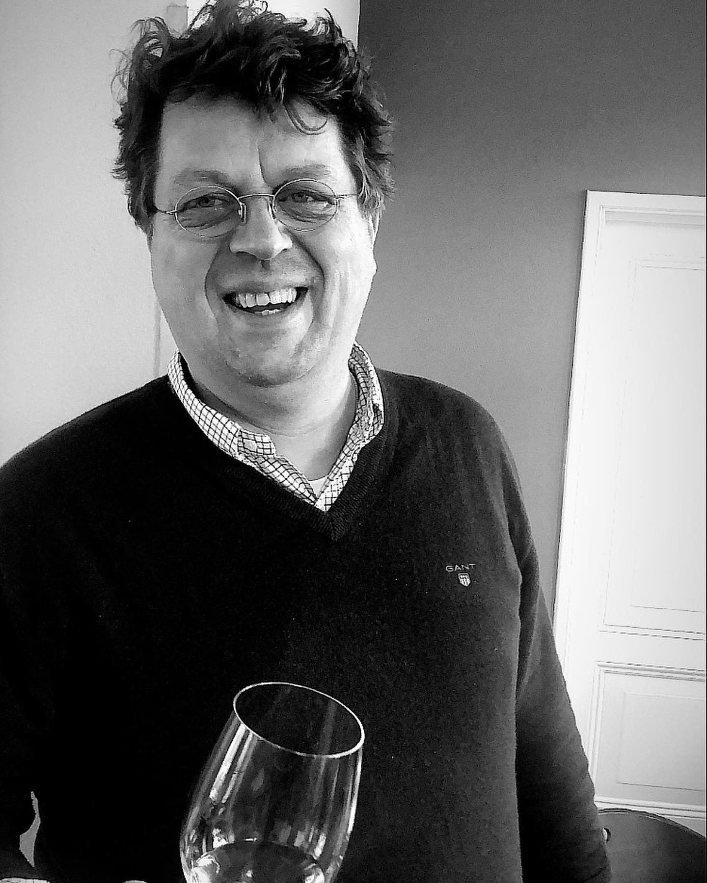Een glimlachende man met een wijnglas in zijn hand. Hij draagt ​​een bril, een donkere trui en een overhemd met een opvallende kraag.