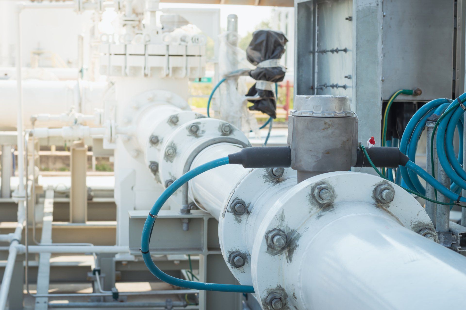White industrial pipes with bolted flanges and attached blue electrical cables. Outdoor setting.
