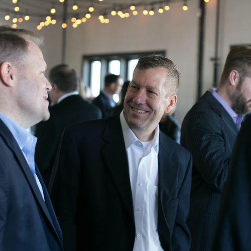 Two men in suits smiling and talking at an event, other people in the background.