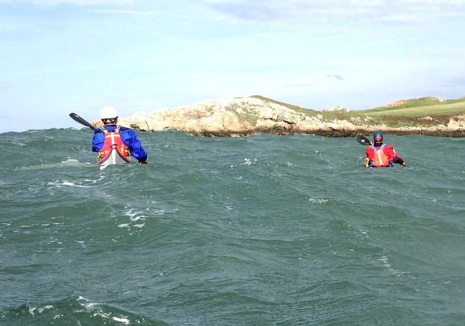 Two kayakers paddling on choppy water near a rocky coastline.
