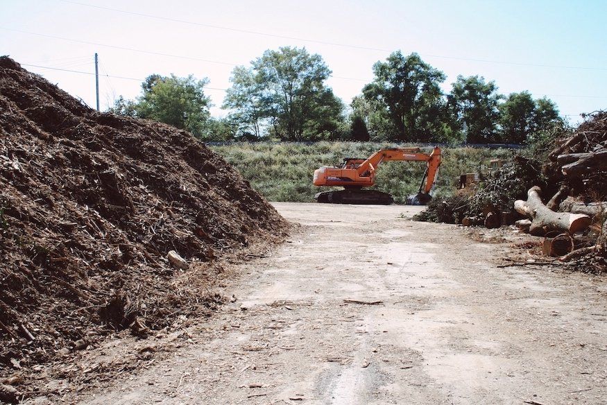 Orange excavator on a dirt lot beside a large woodpile under a clear sky