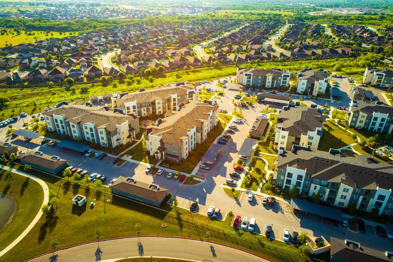 Aerial view of a modern suburban apartment complex with roads, parked cars, and green lawns under bright sunlight