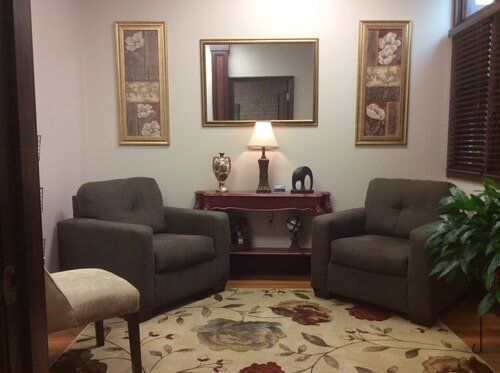 Cozy waiting room with two brown armchairs, red table, and floral rug. Mirror and artwork on the walls.