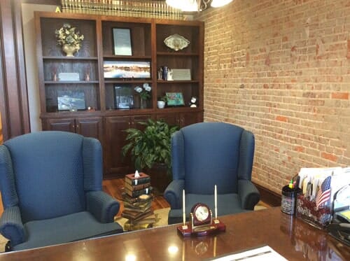 Office with two blue armchairs facing a desk. Bookshelves, brick wall, potted plant.