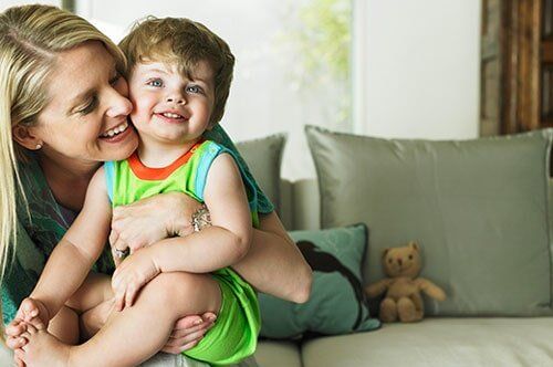 Woman holding and kissing a smiling child indoors on a couch.