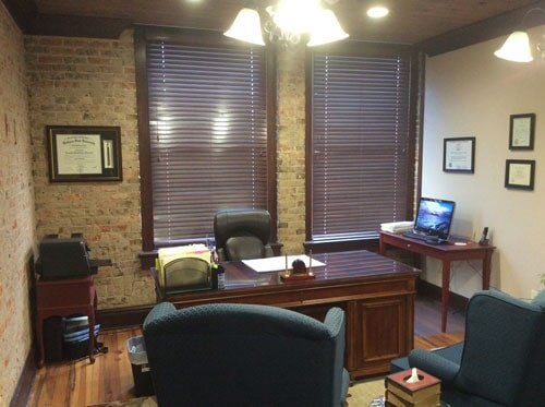 Office with exposed brick wall, desk, windows with blinds, and chairs. Laptop on small table.