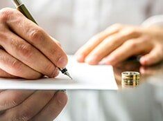 Person writing with a pen on white paper, wedding ring beside the hand on a reflective surface.