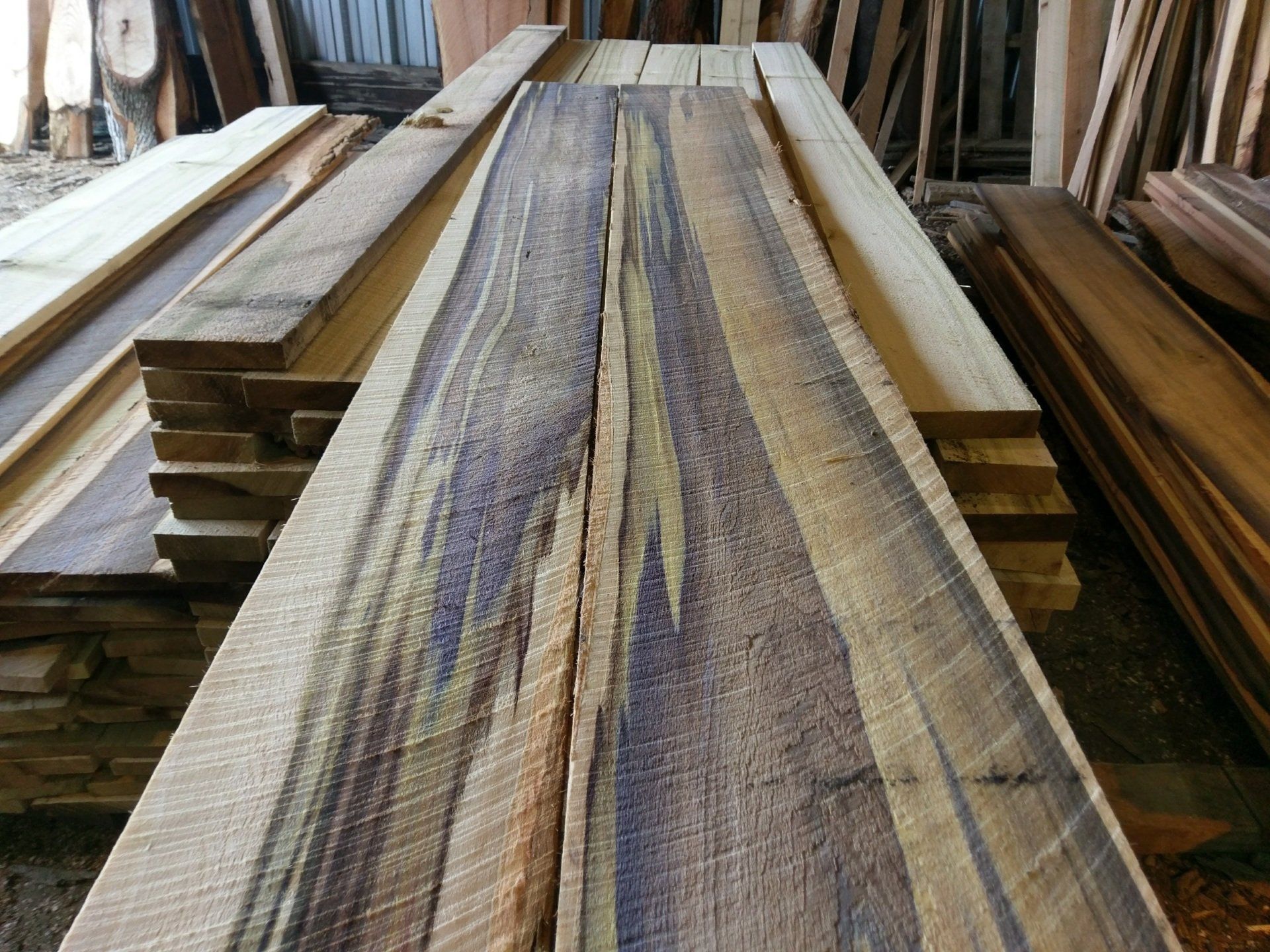 Stacked wooden planks in a workshop, including long boards with a purple-tinted grain on top.