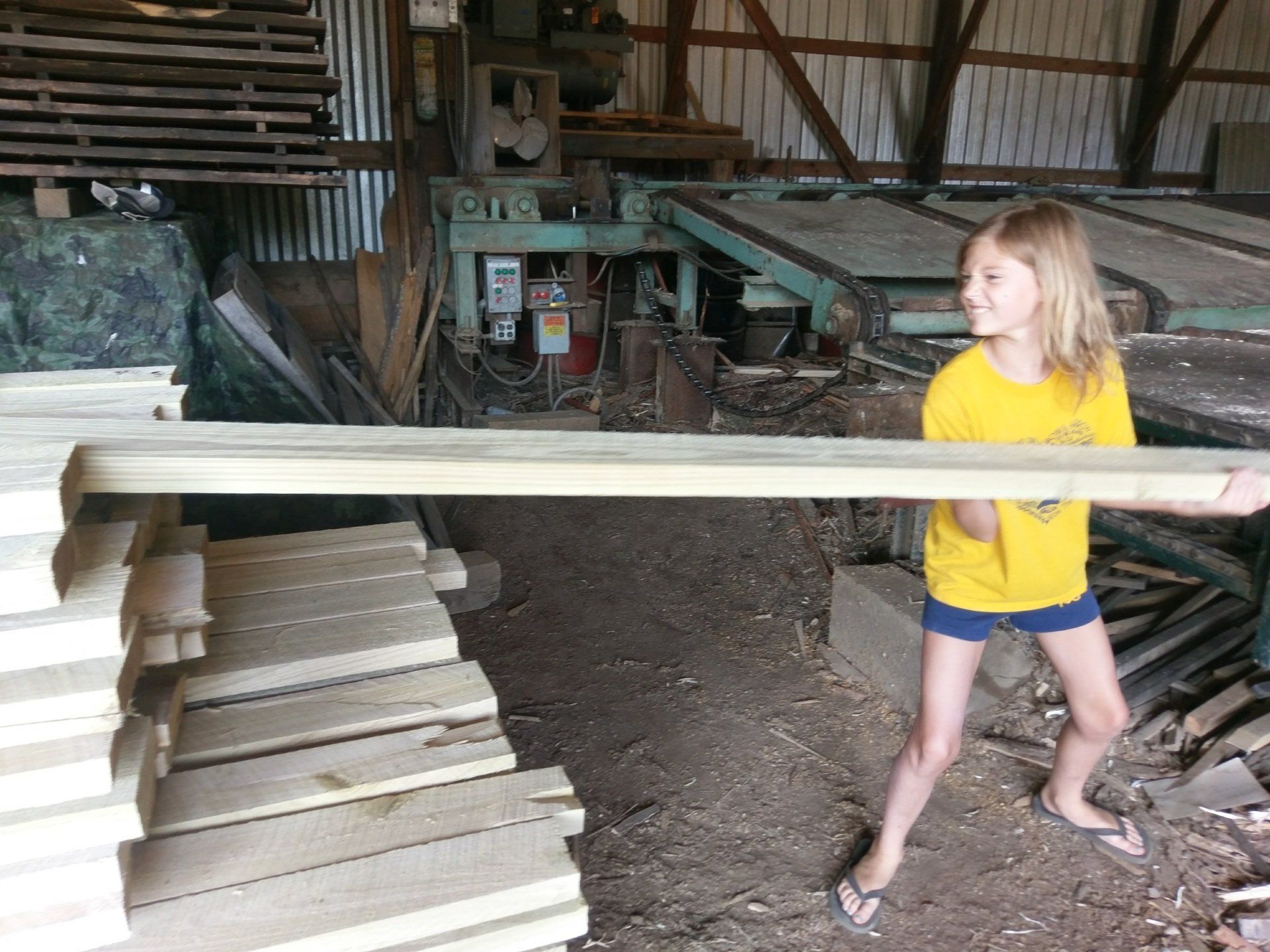 Child in a yellow shirt carrying a long board in a woodshop, with stacks of lumber nearby.