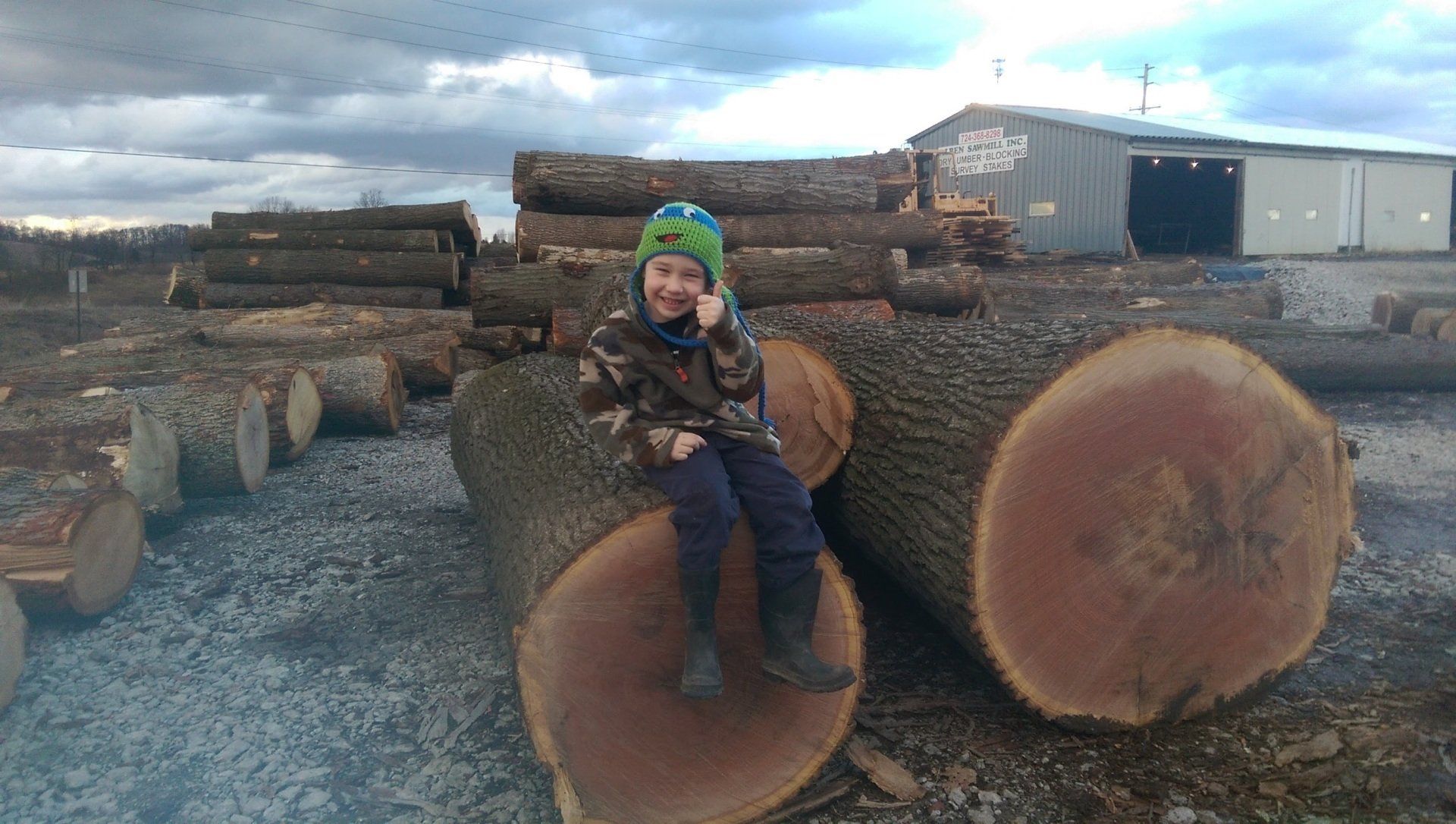 Child sitting on stacked logs in a lumber yard, wearing a green hat and blue jacket.