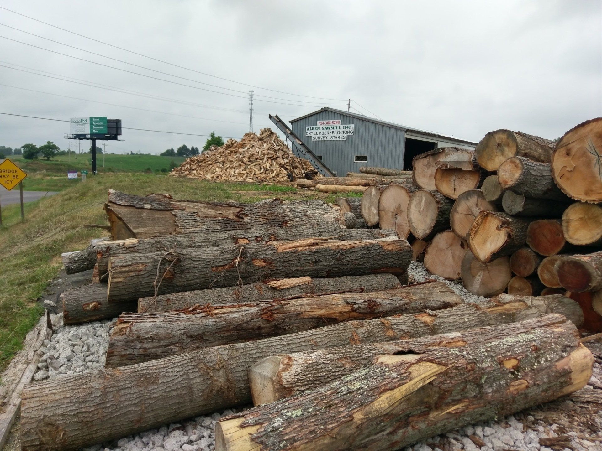 Large stacks of cut logs beside a rural road and a sawmill building under cloudy skies