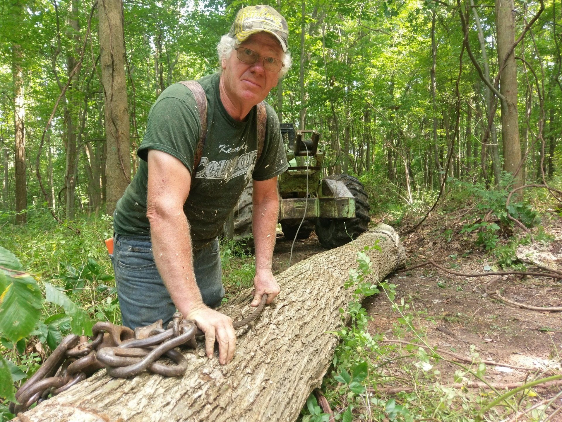 Man standing on a large fallen log in a wooded trail, with an off-road vehicle behind him.