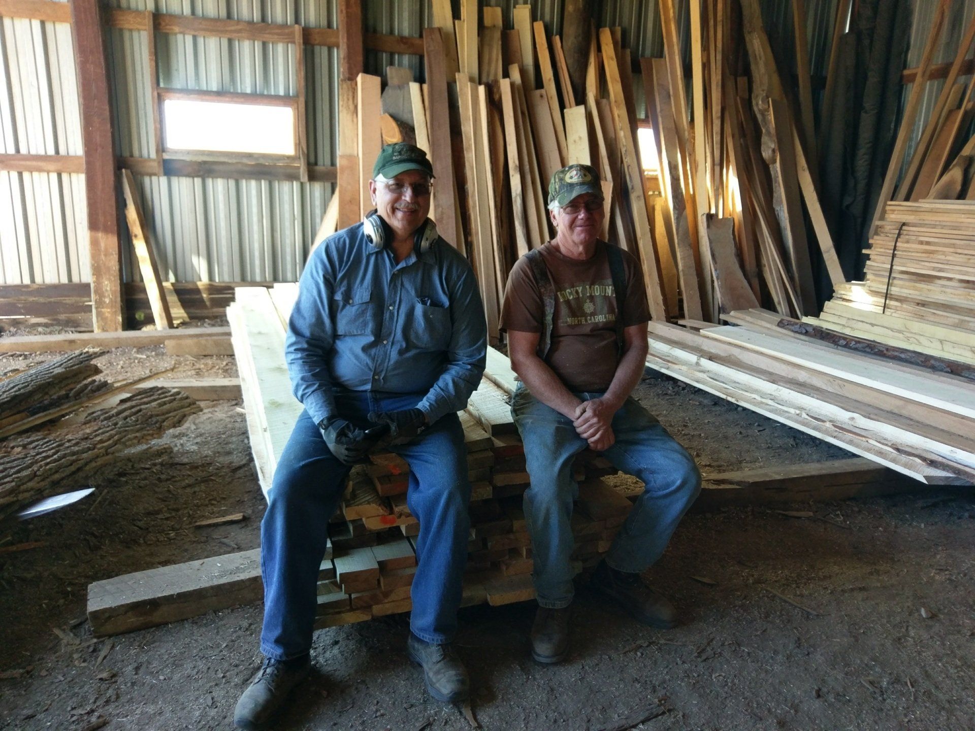 Two people sitting on lumber stacks inside a woodshop, with boards stacked along the walls.