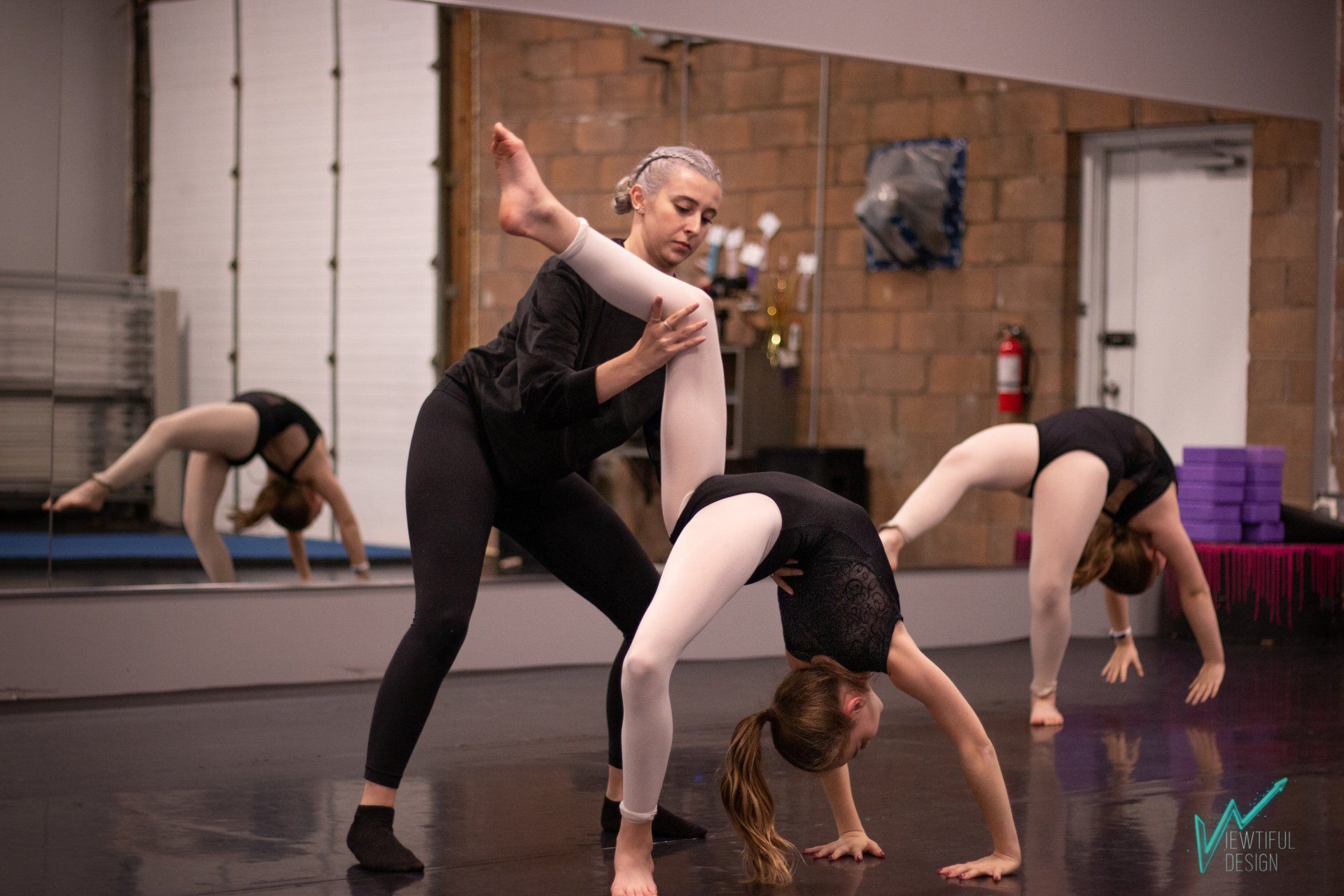 Dancers in black practice a contortion pose in a studio with mirrors and a wooden floor.
