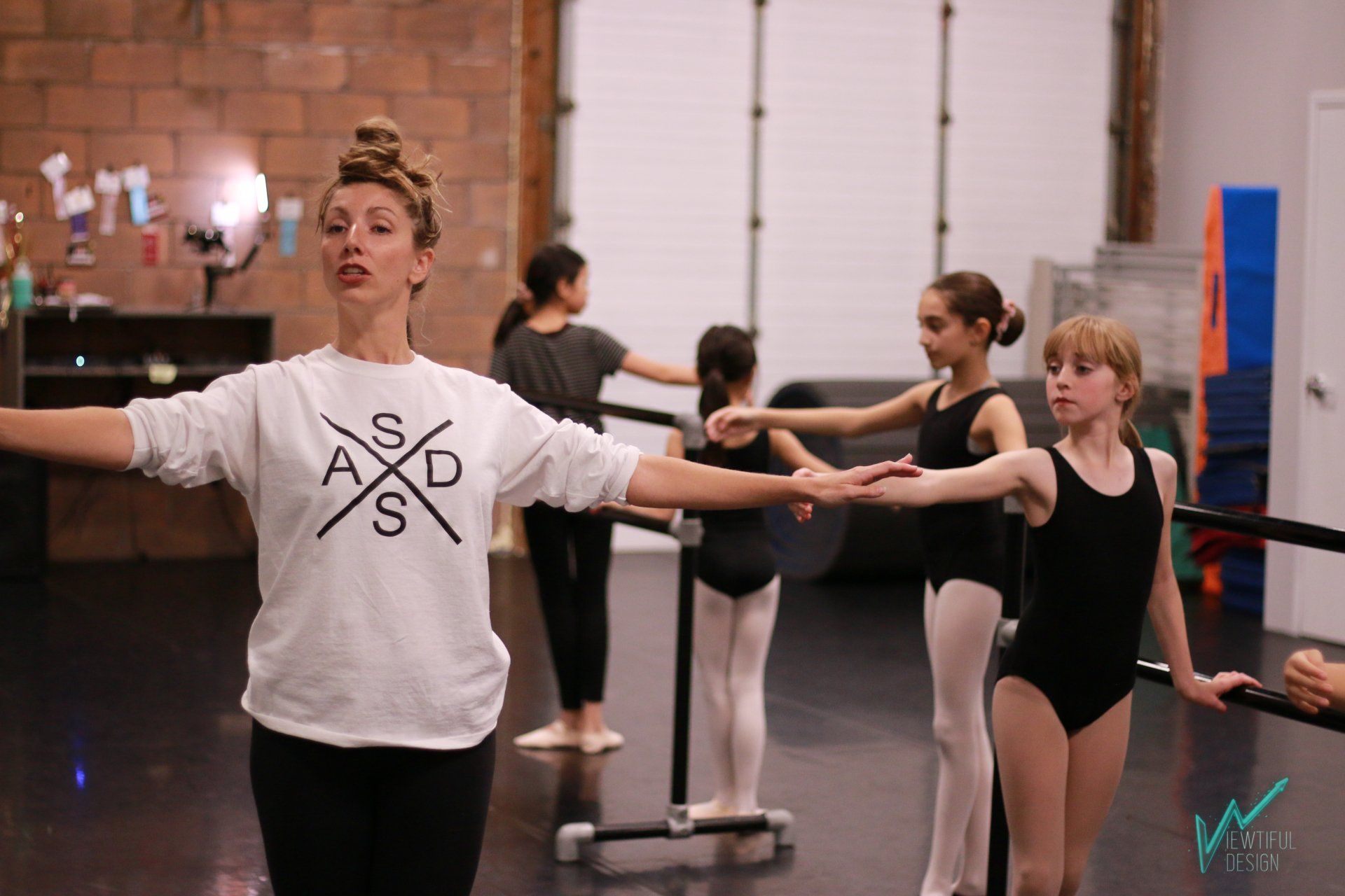 Dance instructor leading ballet students at the barre in a studio, arms extended during practice.