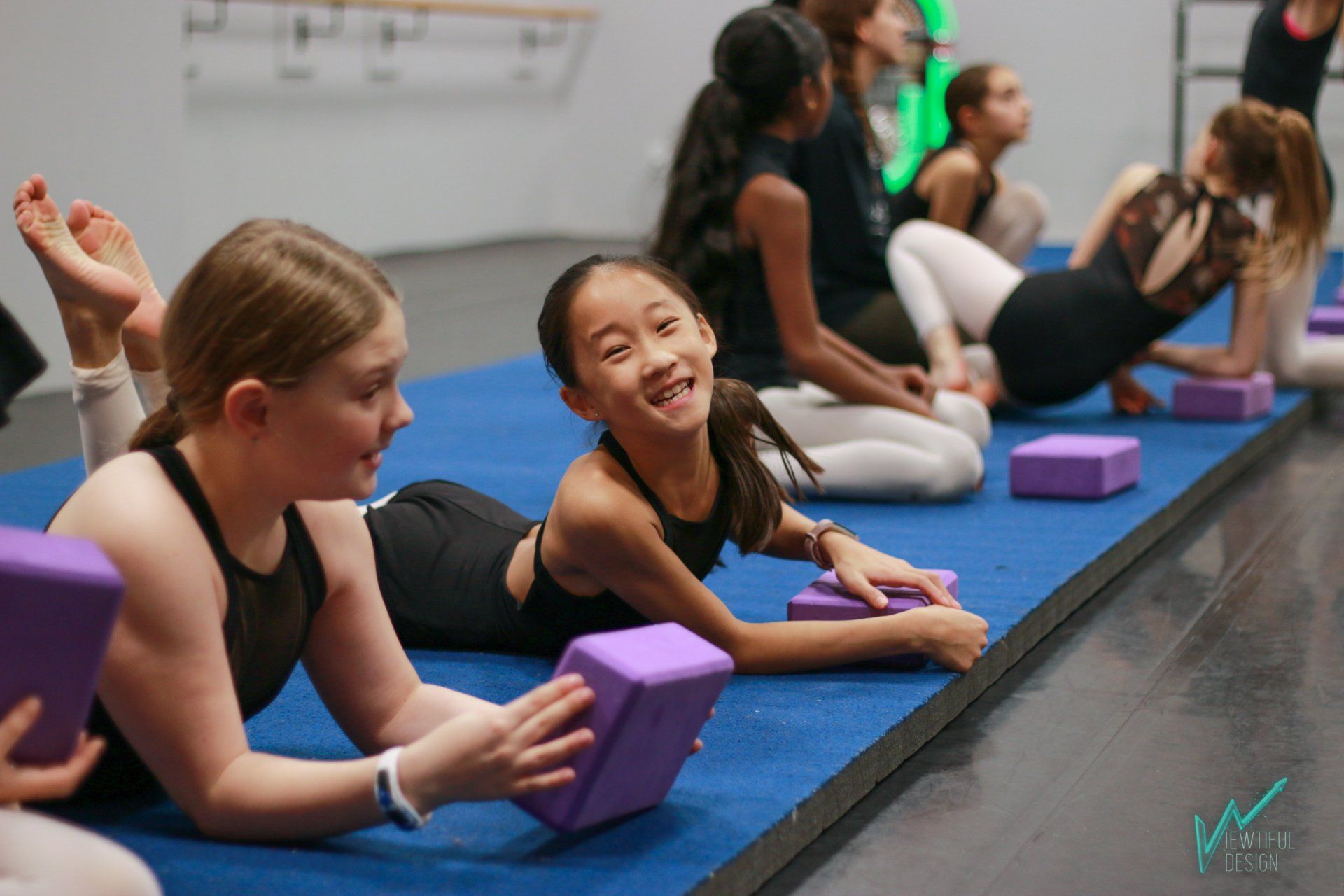 People doing yoga on blue mats in a studio, using purple blocks and smiling during a class