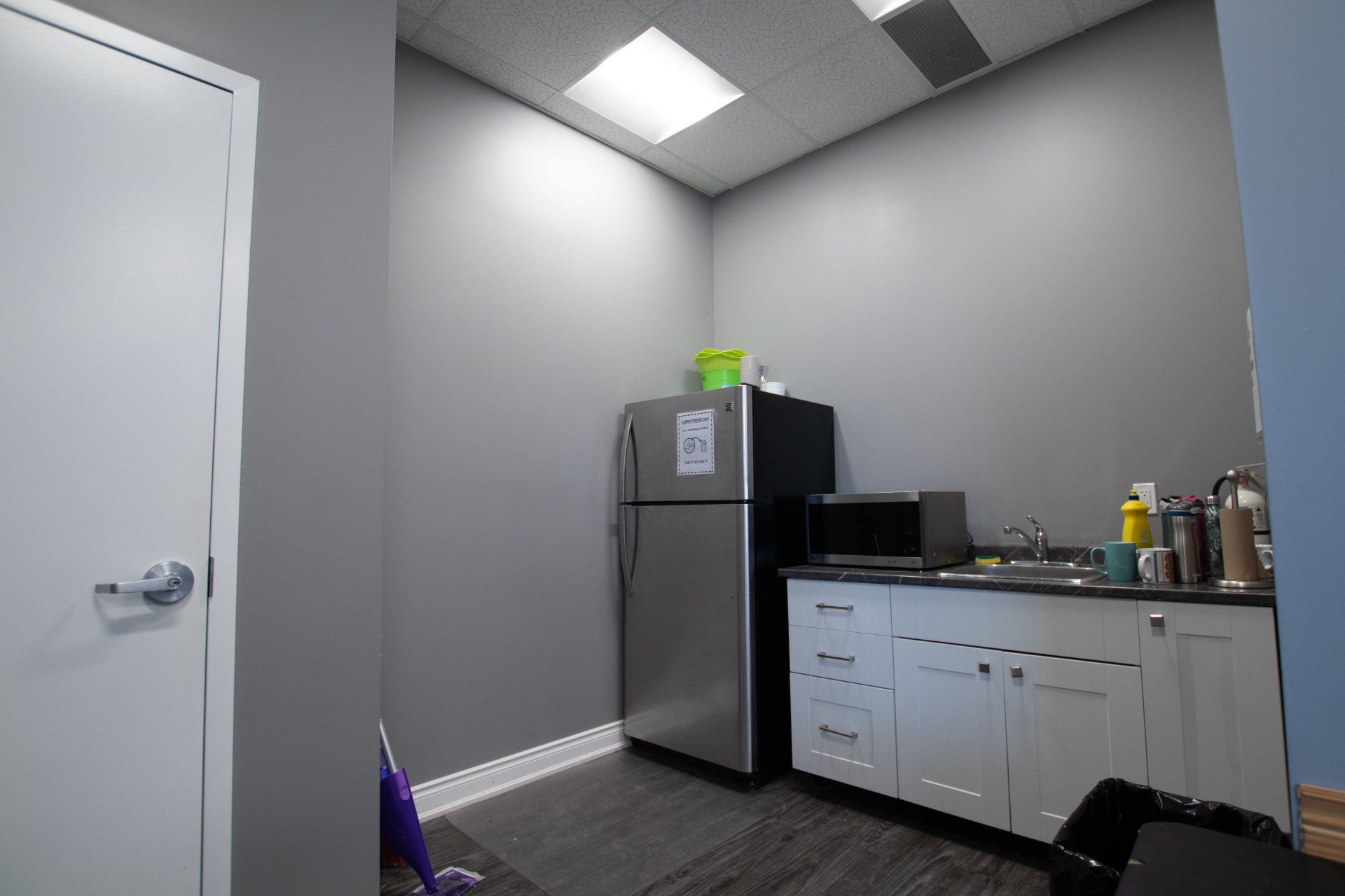 Small break room with fridge, white cabinets, sink, and a closed door under bright ceiling lights.