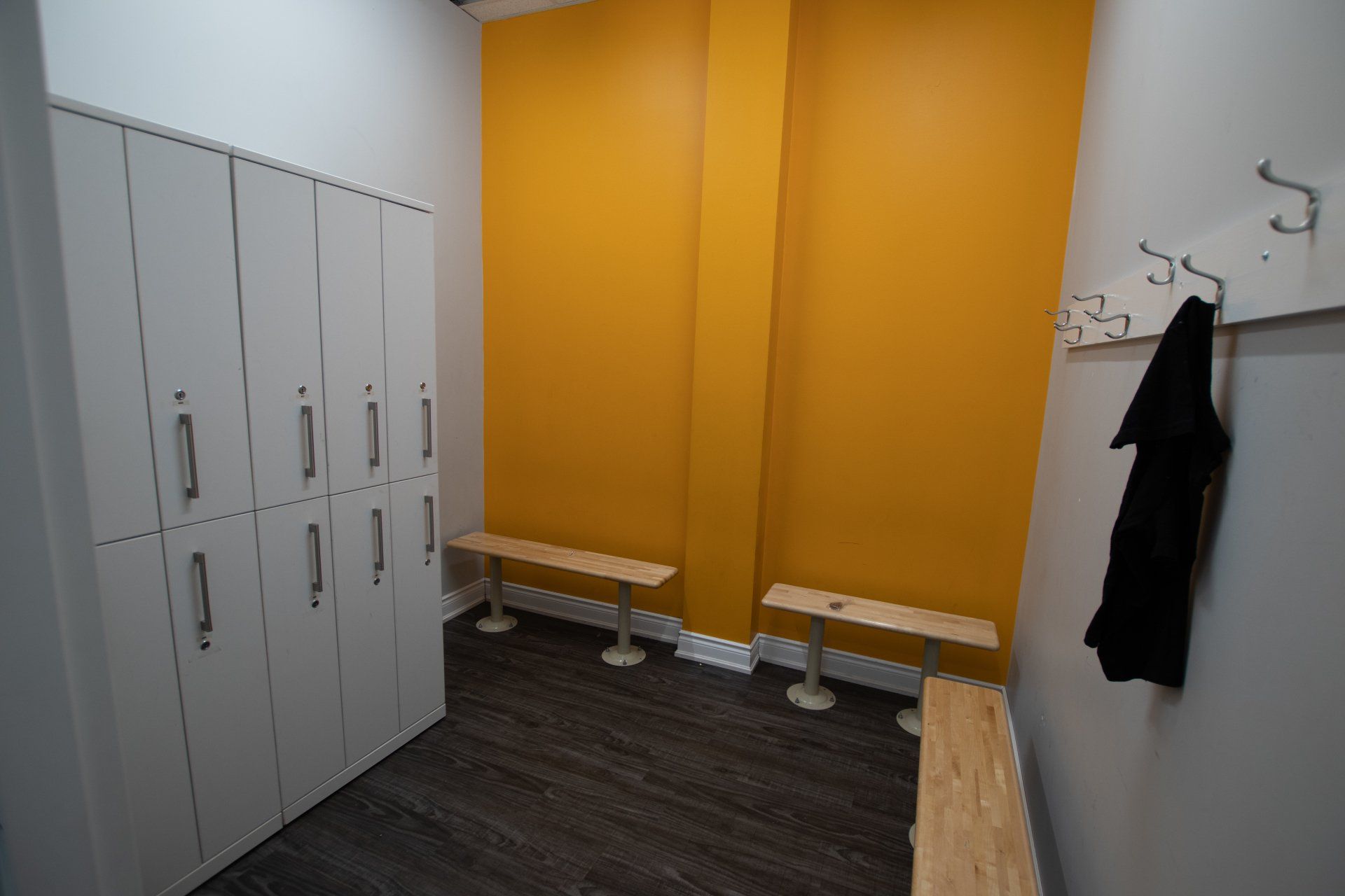Empty locker room with white lockers, orange wall, benches, and coat hooks.