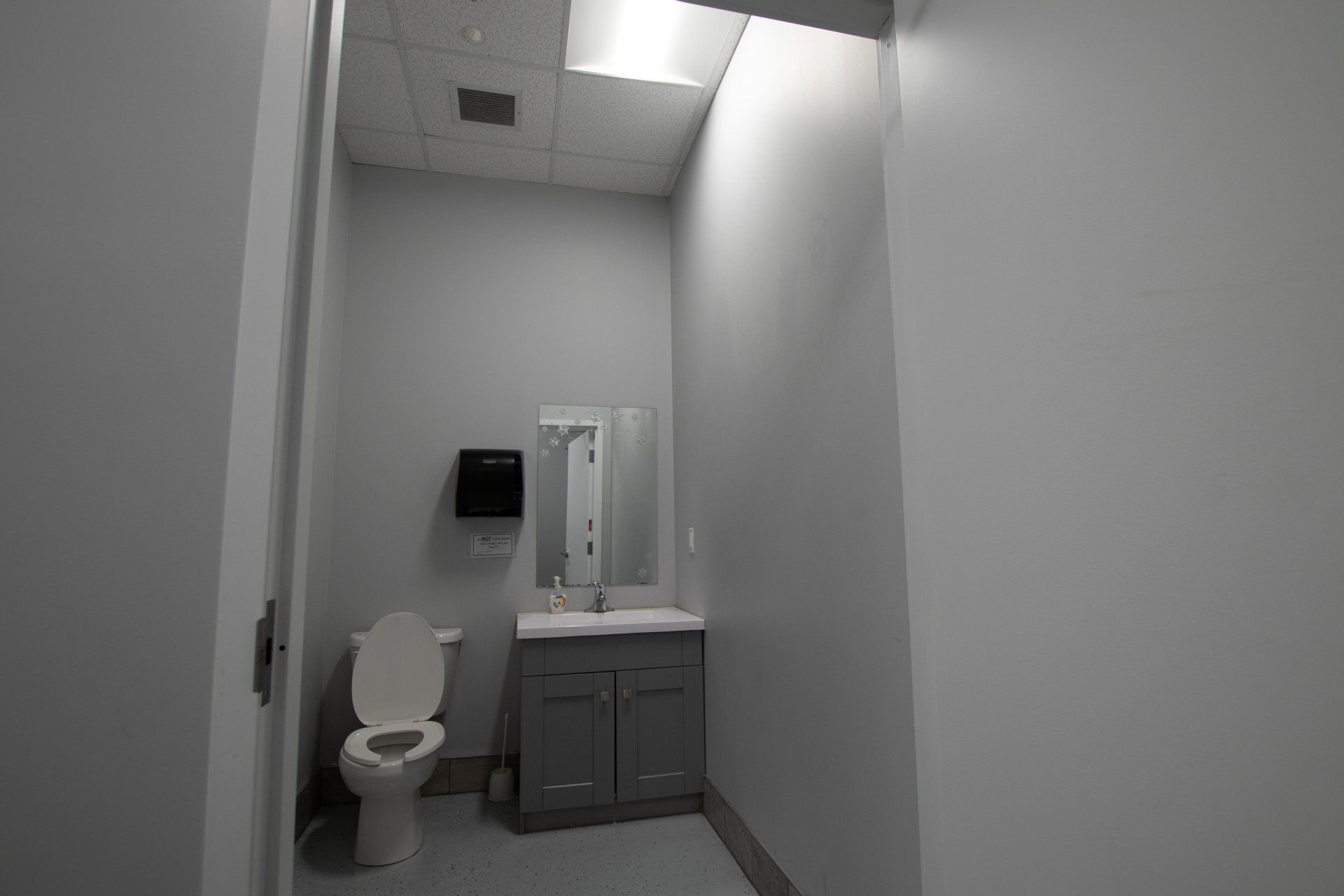 Narrow gray restroom with toilet, sink, mirror, and partially open ceiling tiles.