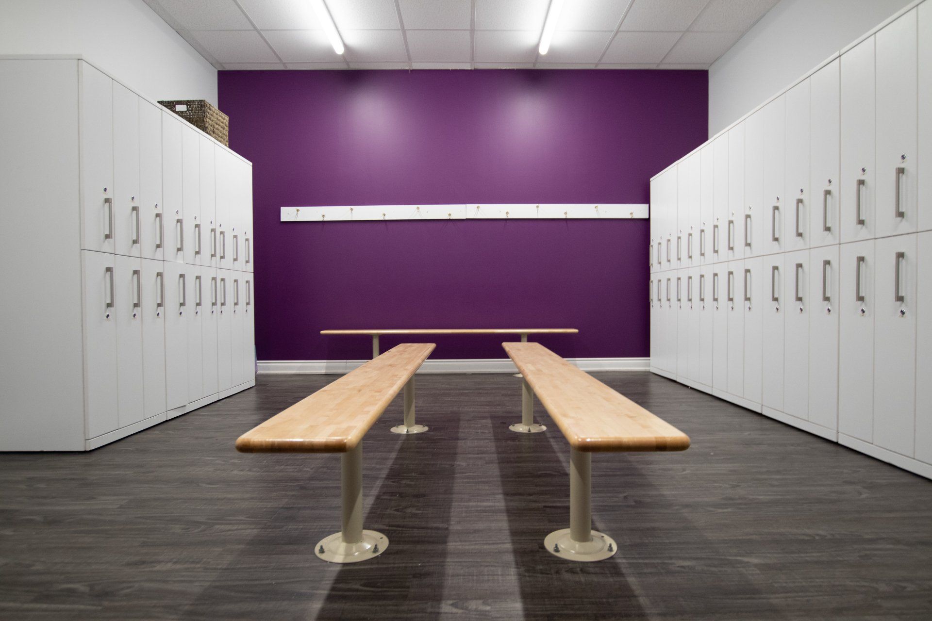 Locker room with white lockers, purple wall, and two wooden benches on a dark floor.