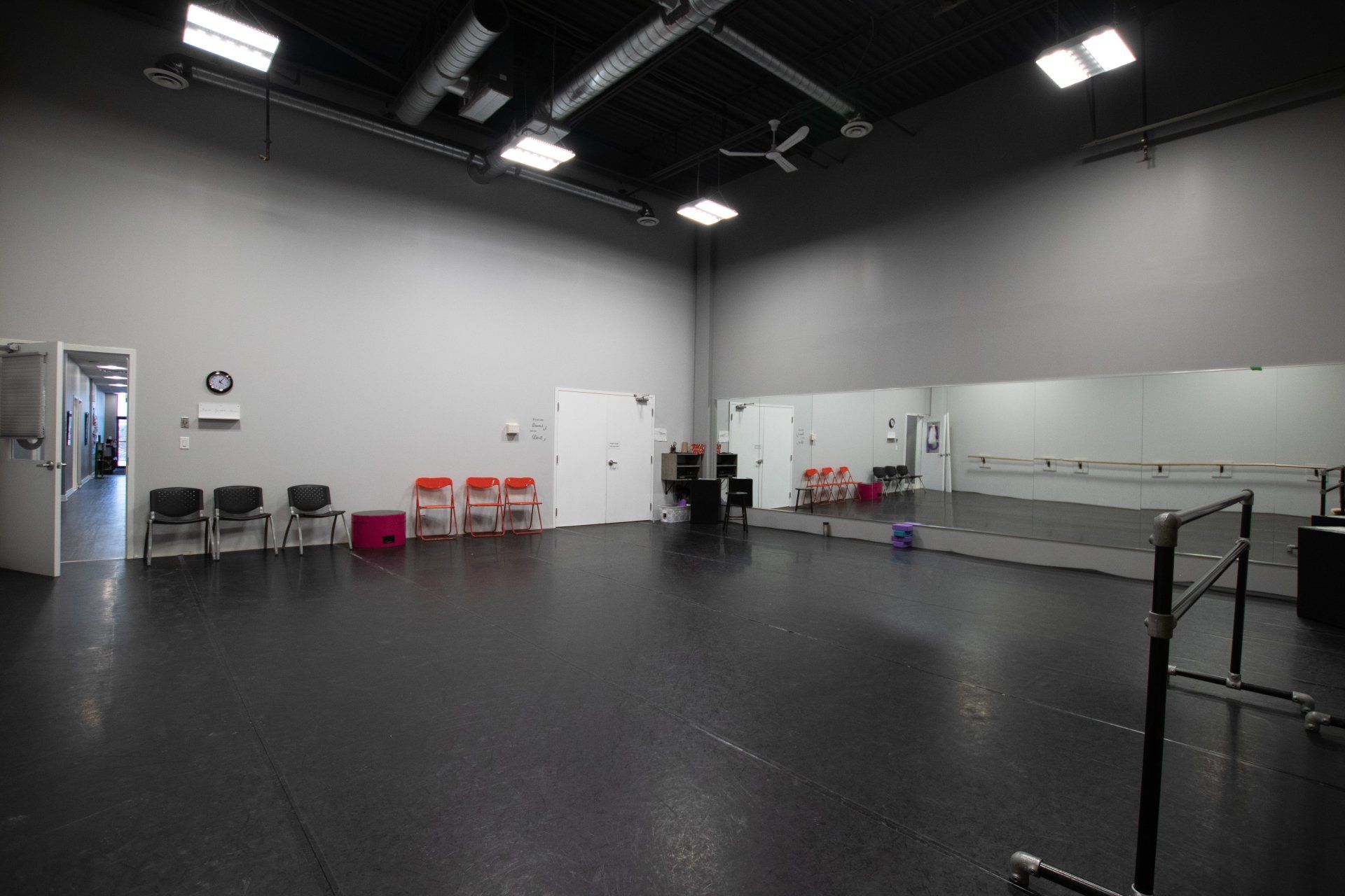 Empty dance studio with black floor, mirrors, ballet barre, chairs, and bright ceiling lights