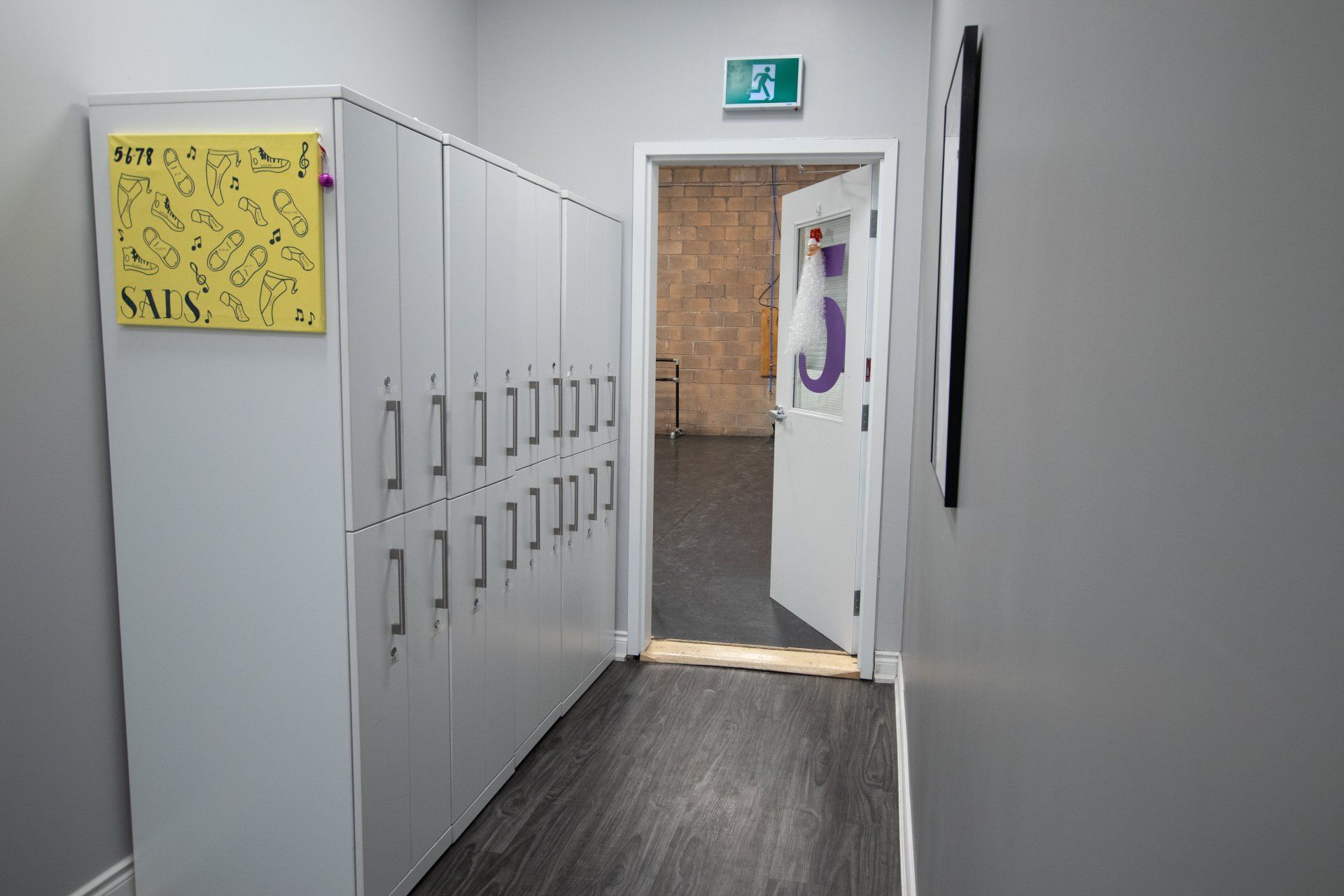 Hallway with gray lockers, a yellow sign, and an open door leading to another room