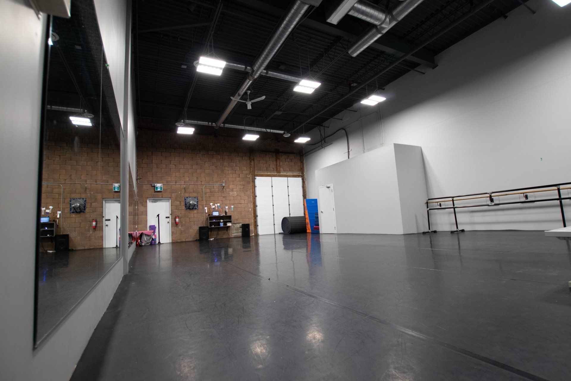 Empty dance studio with gray floor, brick wall, mirrors, and white partition walls under ceiling lights