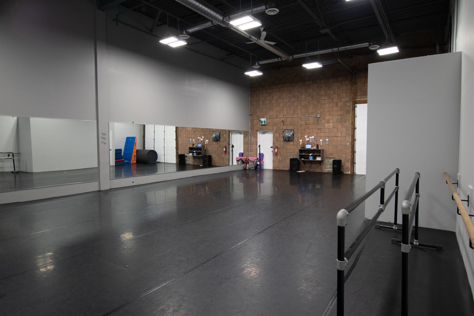 Empty dance studio with mirrored wall, black floor, exposed brick, and ballet barres along the right side.