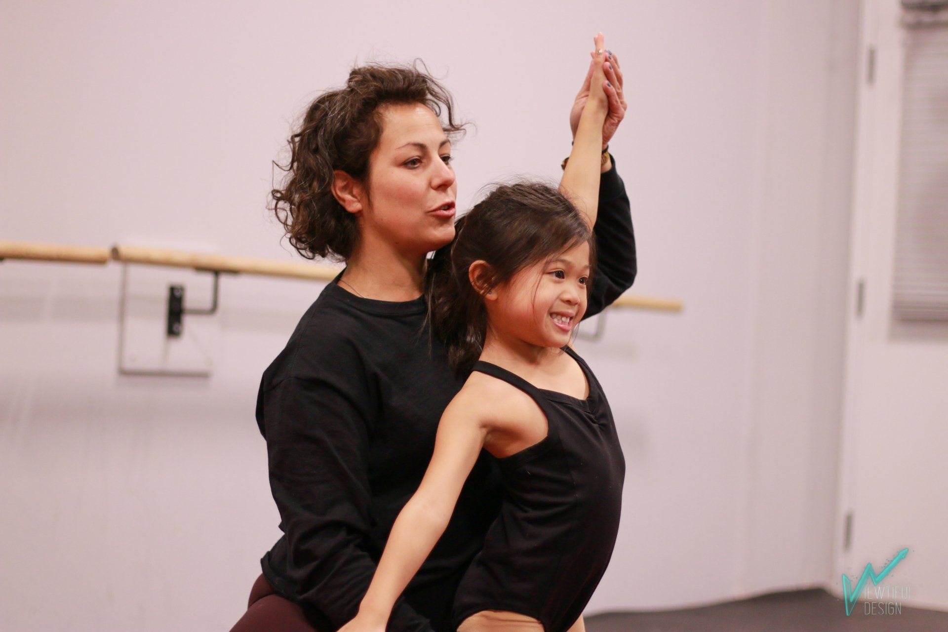 Two dancers in black practice a partnered pose in a studio, one lifting an arm overhead.