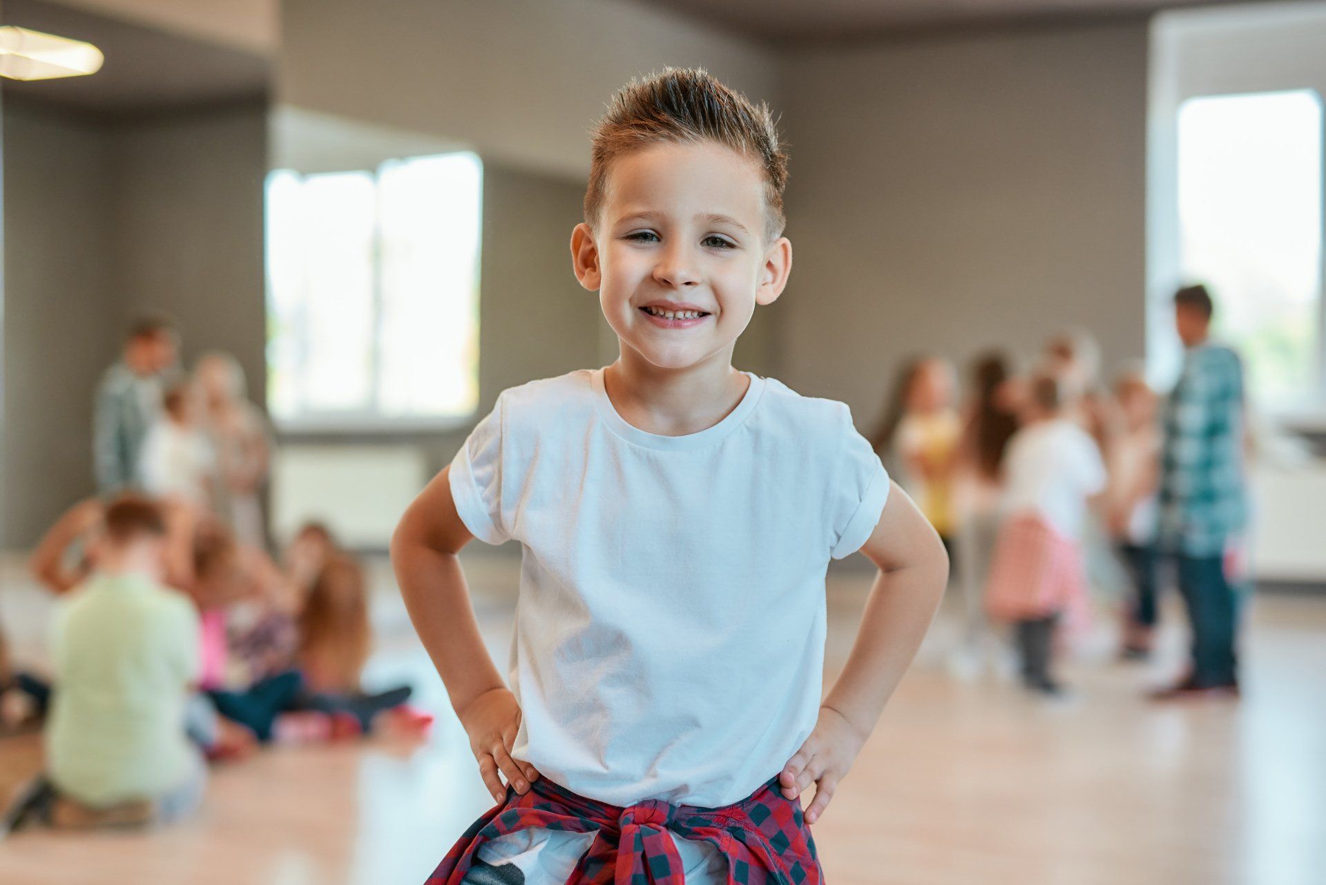 Smiling child in a white shirt standing in a dance studio with others blurred in the background